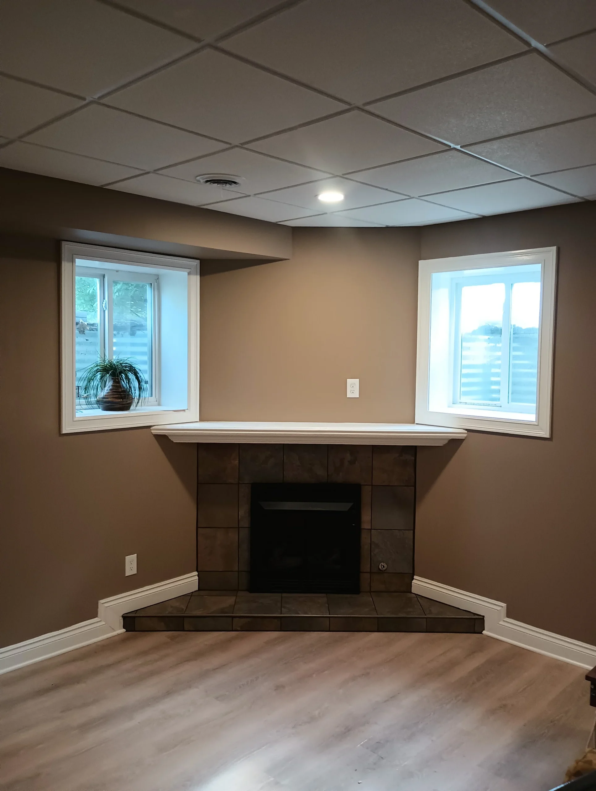 Basement corner with two windows, a small potted plant on the left windowsill, a tiled fireplace below a white mantel, beige walls, light wood flooring, and a white drop ceiling with recessed lighting.