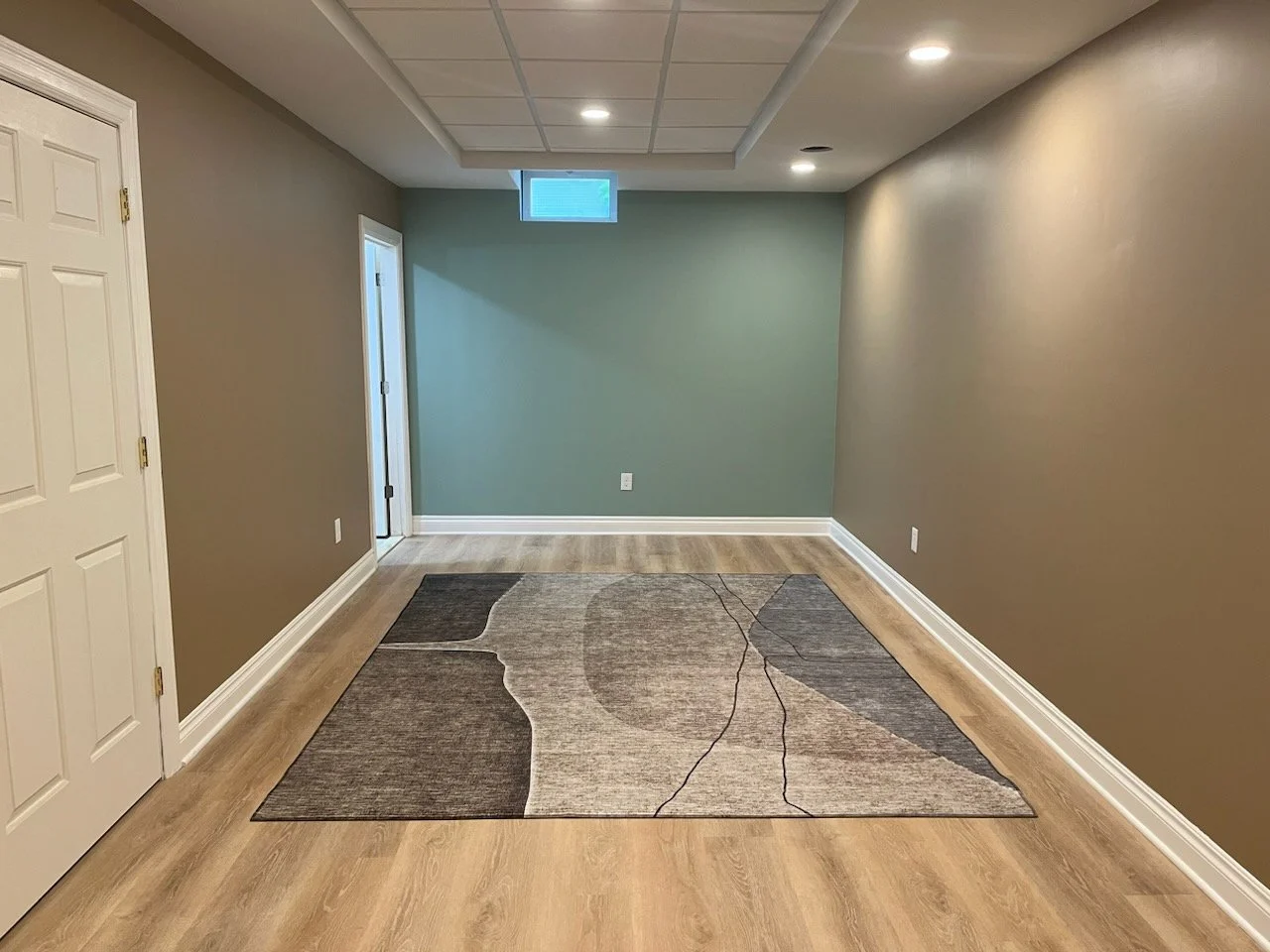 A finished basement room with light wood flooring, beige walls, white trim, recessed ceiling lights, and a modern area rug with abstract patterns in gray tones. A small window and white double doors are visible.