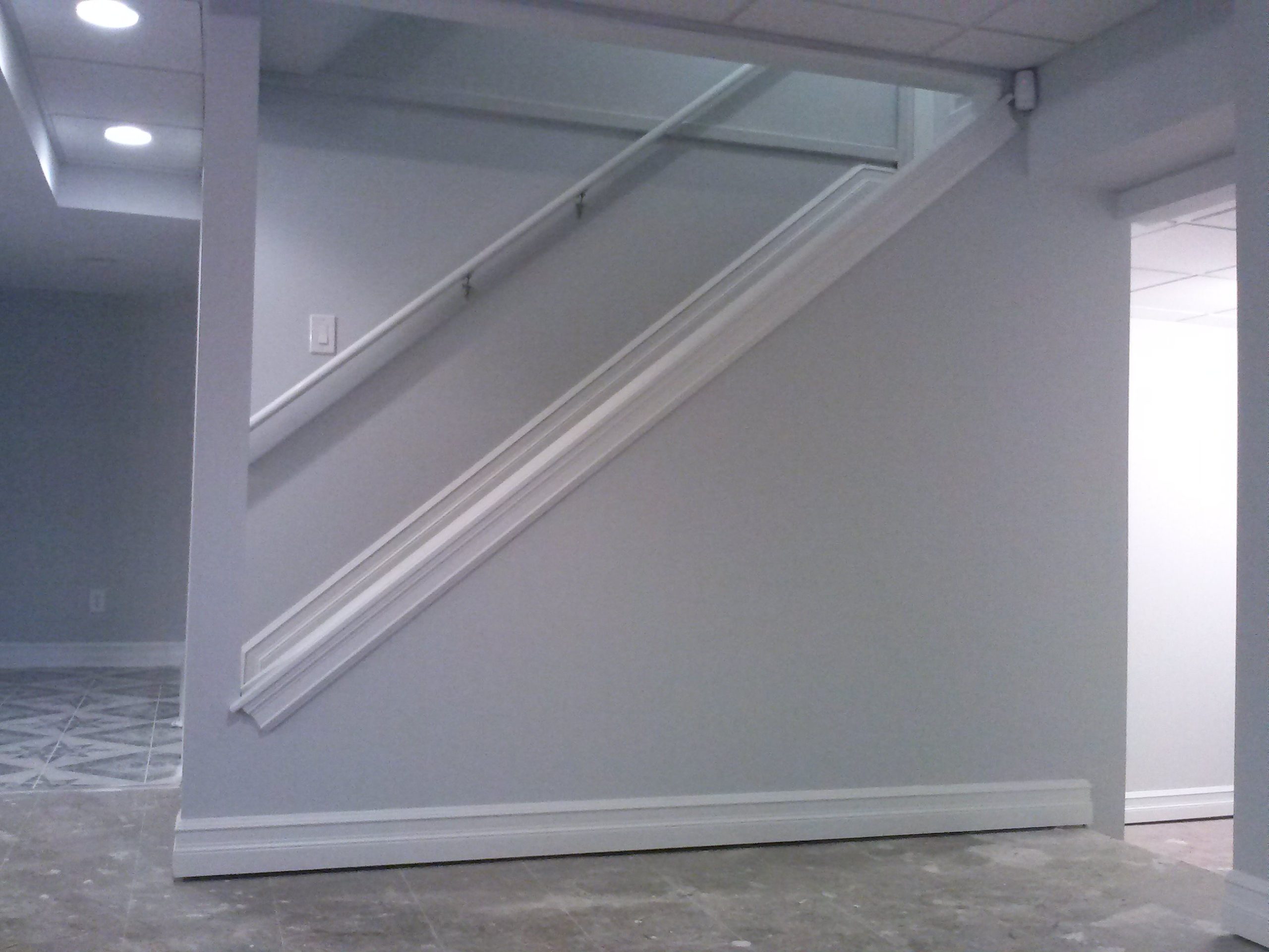 A modern basement with light gray walls and tiled floor, featuring a staircase wall with white trim and a doorway leading to another bright room. Ceiling lights brighten the space.